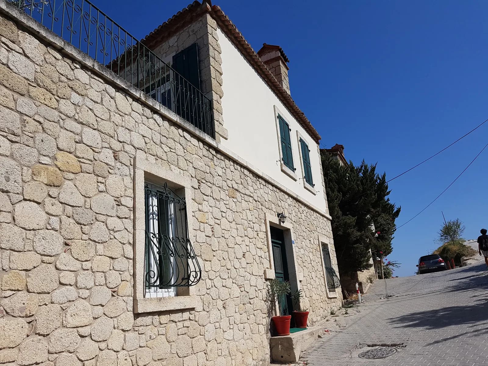 A stone house with green shutters and wrought-iron window grilles on a sloping paved street in Alaçatı