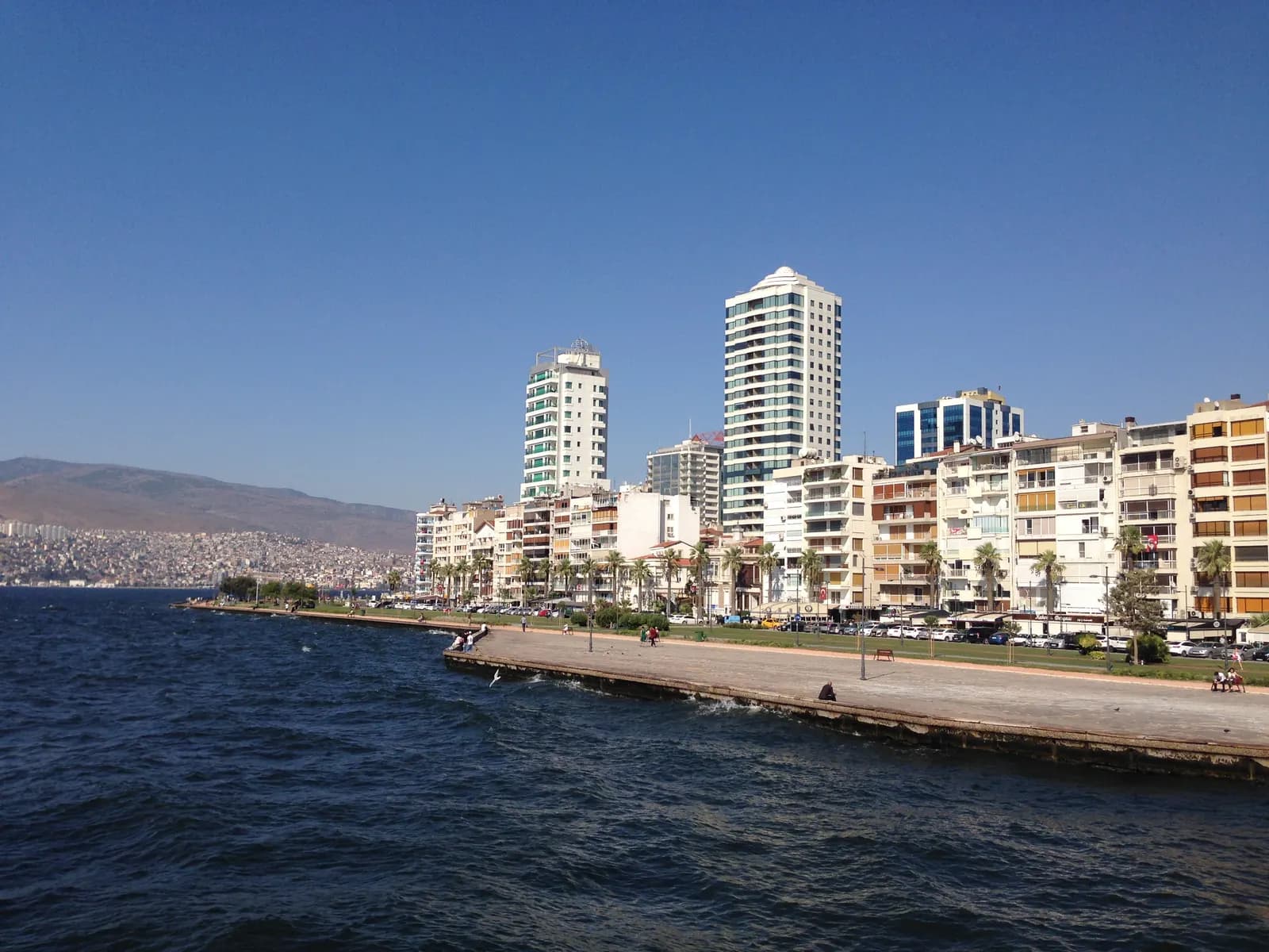 The seawall promenade at the tip of Alsancak with apartment blocks and two towers behind, and the bay on the left