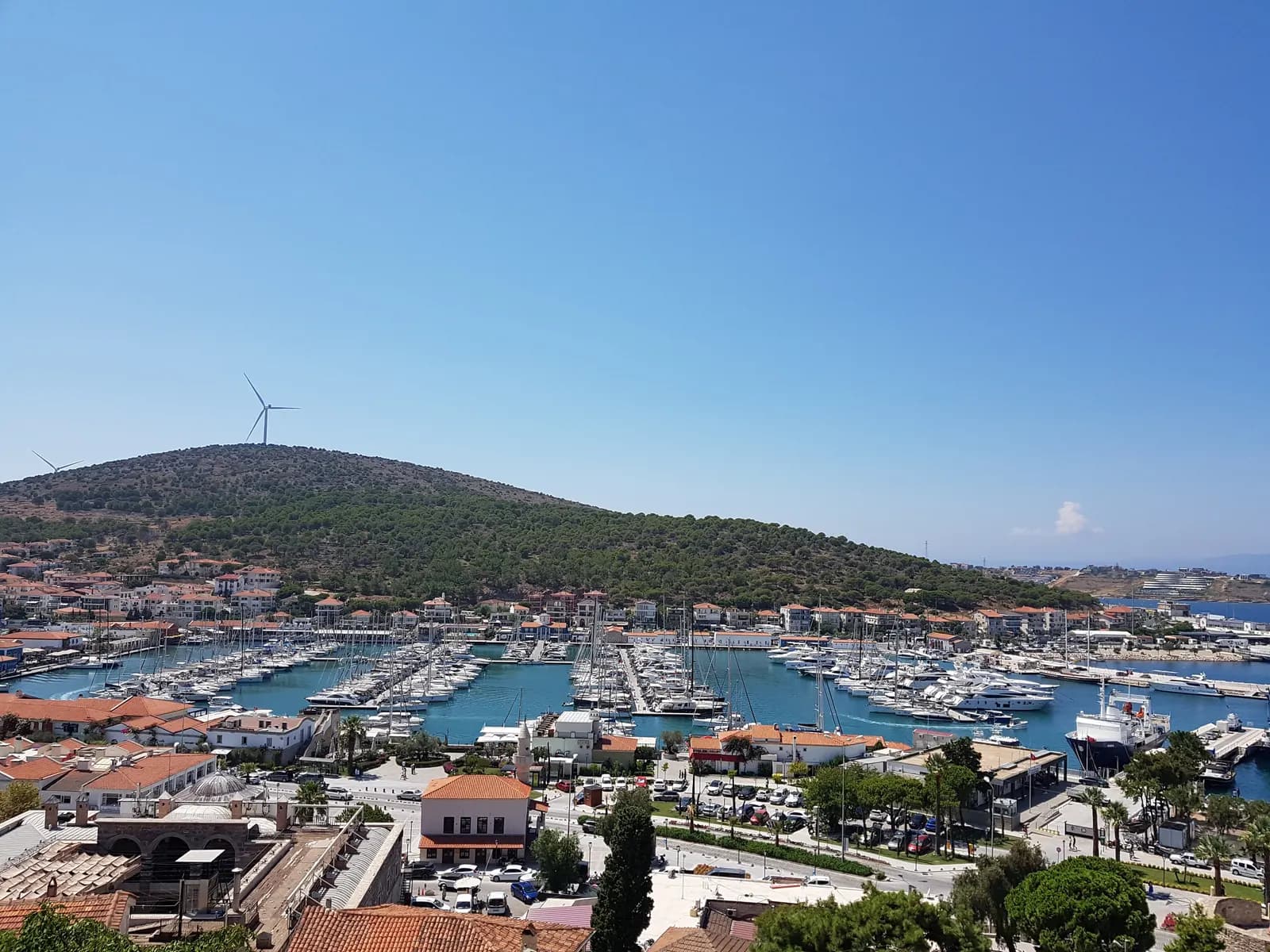 Çeşme marina full of yachts seen from the castle, with tiled roofs in front and a pine-covered hill with a wind turbine behind