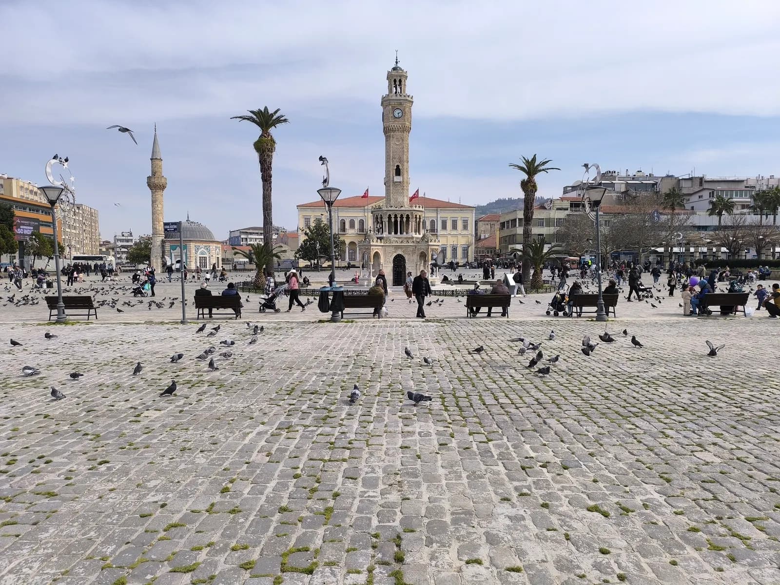 The İzmir clock tower on Konak Square, with a small tiled mosque and its minaret, palm trees, benches and pigeons on the cobbled square