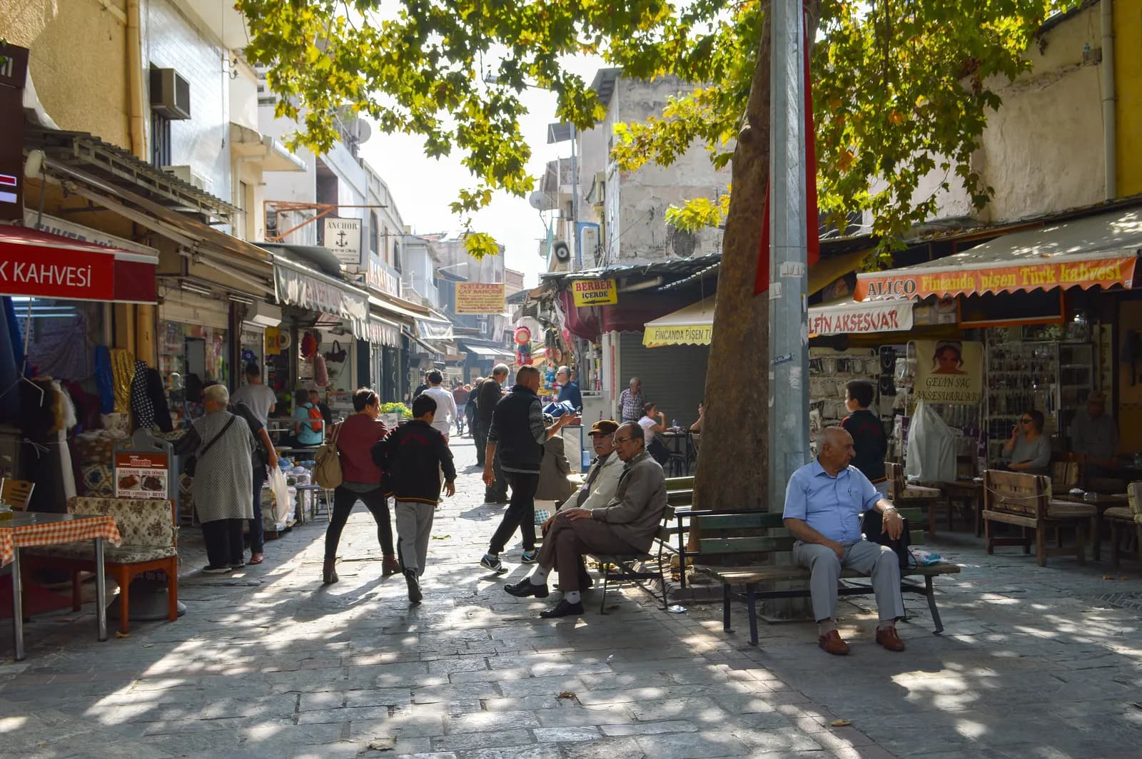 A shaded street in the Kemeraltı market with shops, coffee houses and men sitting on benches under a plane tree