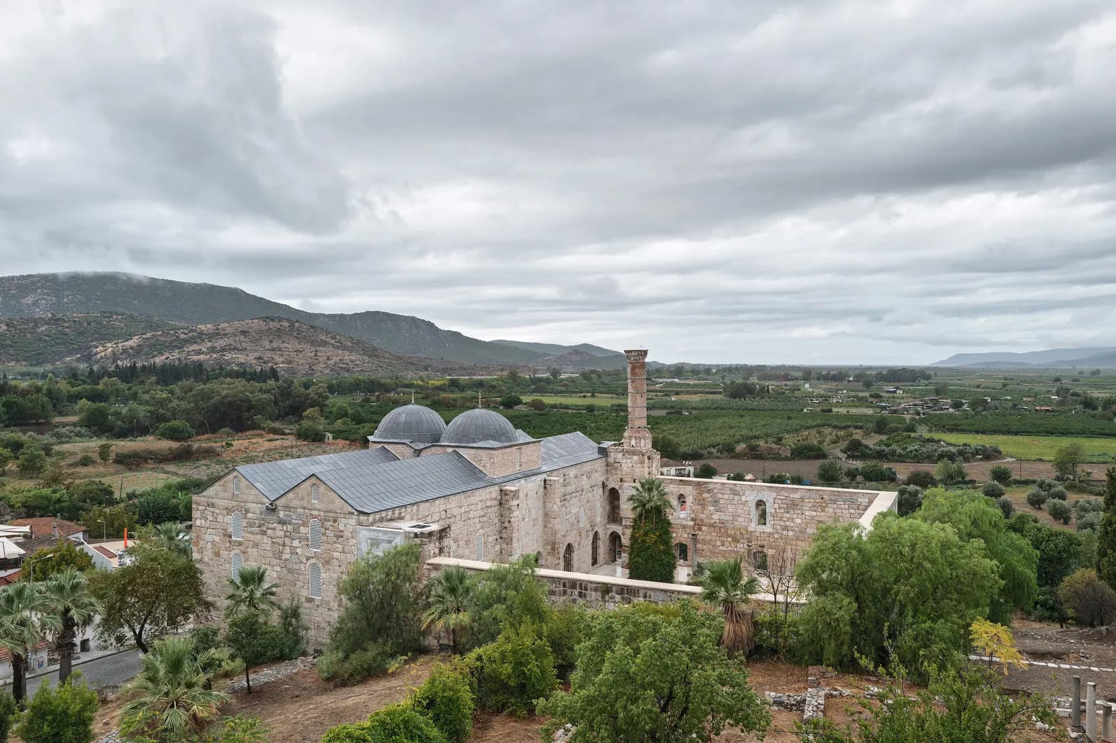 The stone İsa Bey Mosque in Selçuk with its two grey domes and a broken minaret, among trees, with farmland and hills behind