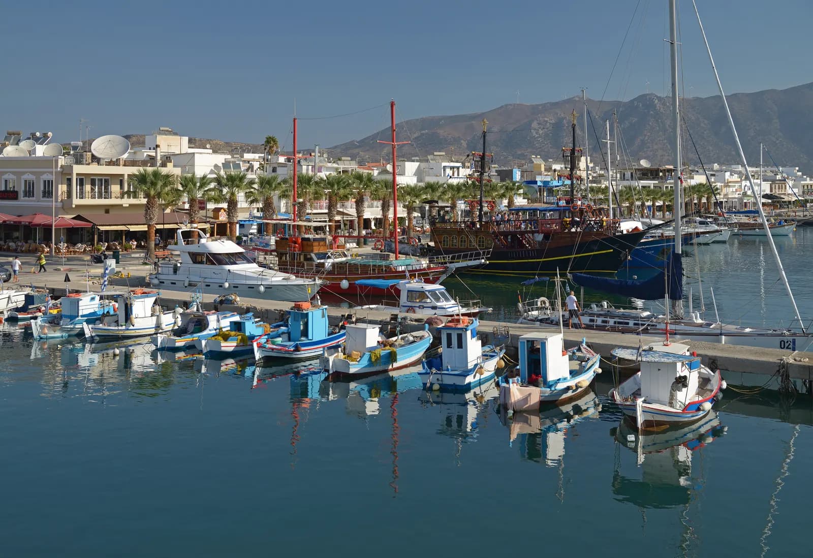 Kardamena harbour with small fishing boats and excursion boats at the quay, waterfront cafes under palm trees and mountains behind