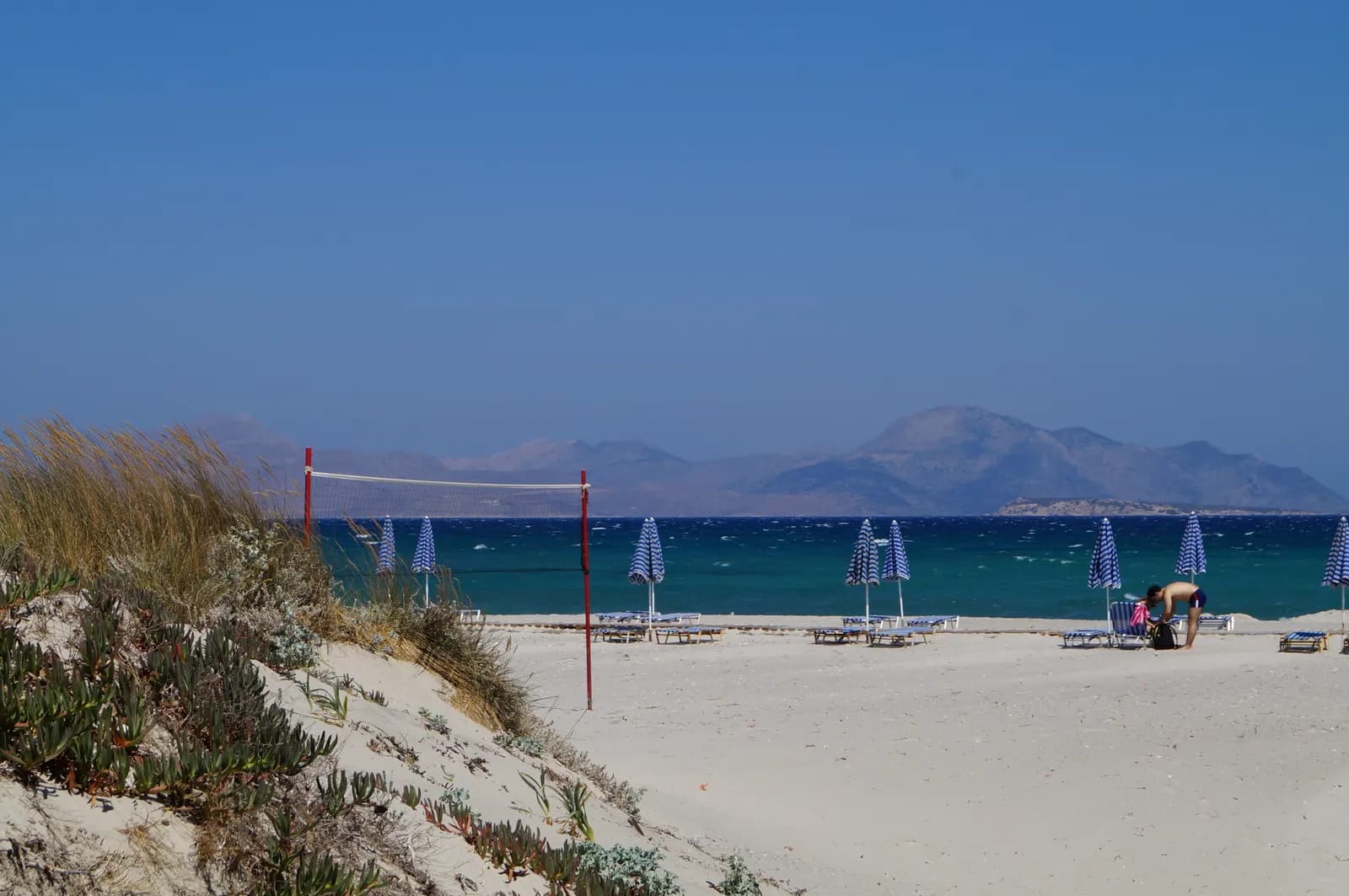 Sand dunes with beach grass above Marmari beach, striped umbrellas and sunbeds by a turquoise sea, and island mountains across the water