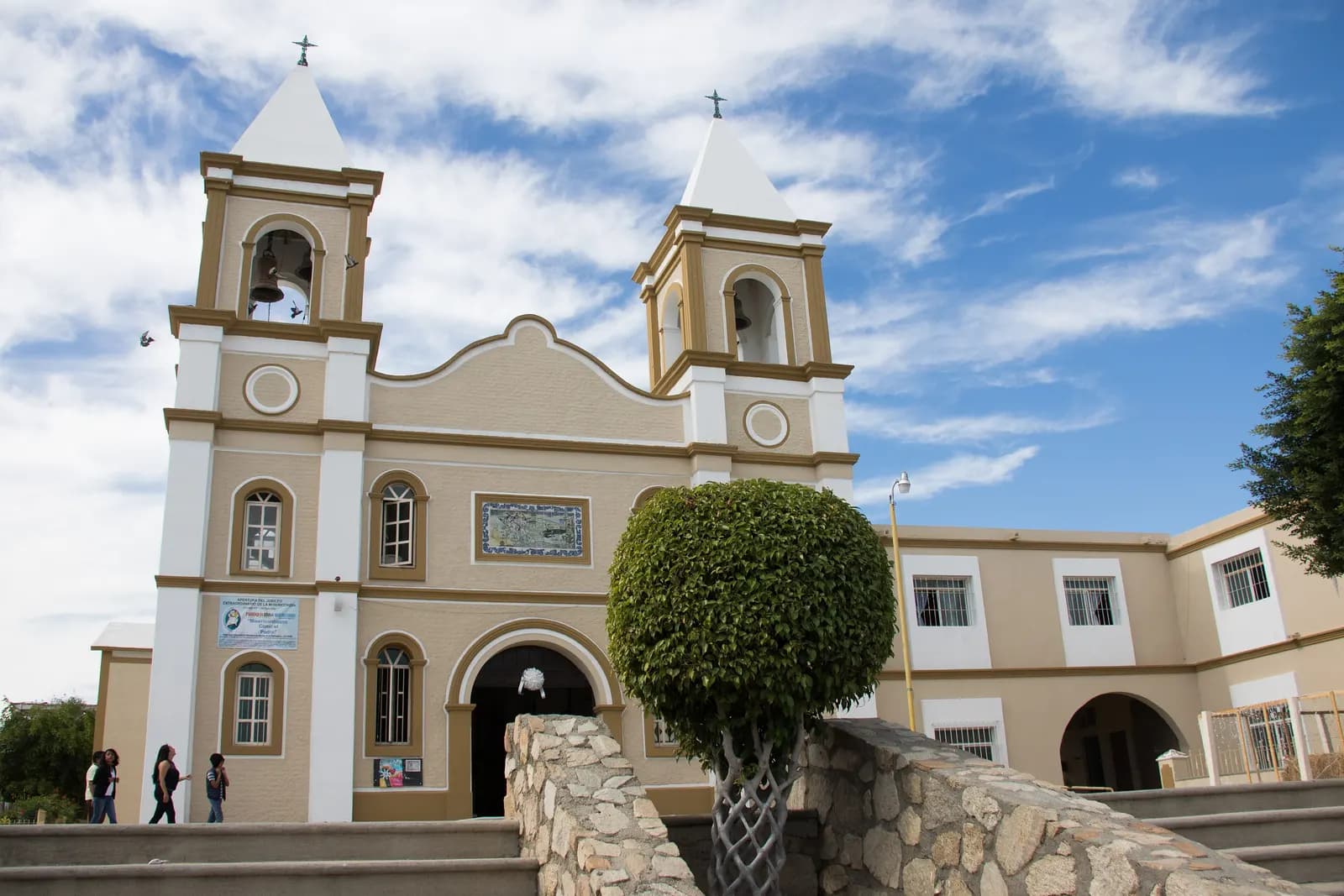 The twin bell towers of the mission church in San José del Cabo, with a tiled mural on the facade and people walking past the steps