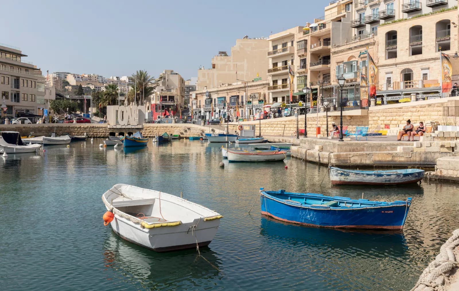 Small wooden boats moored in Spinola Bay in St Julian's, with the stone quay, cafés and apartment buildings along the waterfront