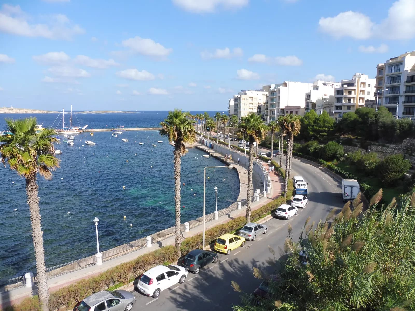 The palm-lined seafront road in St Paul's Bay, with cars parked along the kerb, apartment blocks on the right and boats moored in the bay