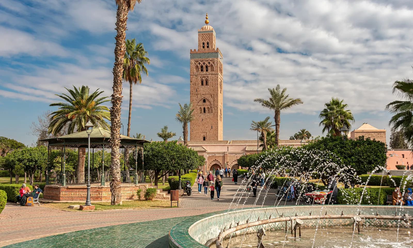 The Koutoubia minaret rising above palm trees and the garden beside it, with a tiled fountain, a bandstand and people walking in the sun