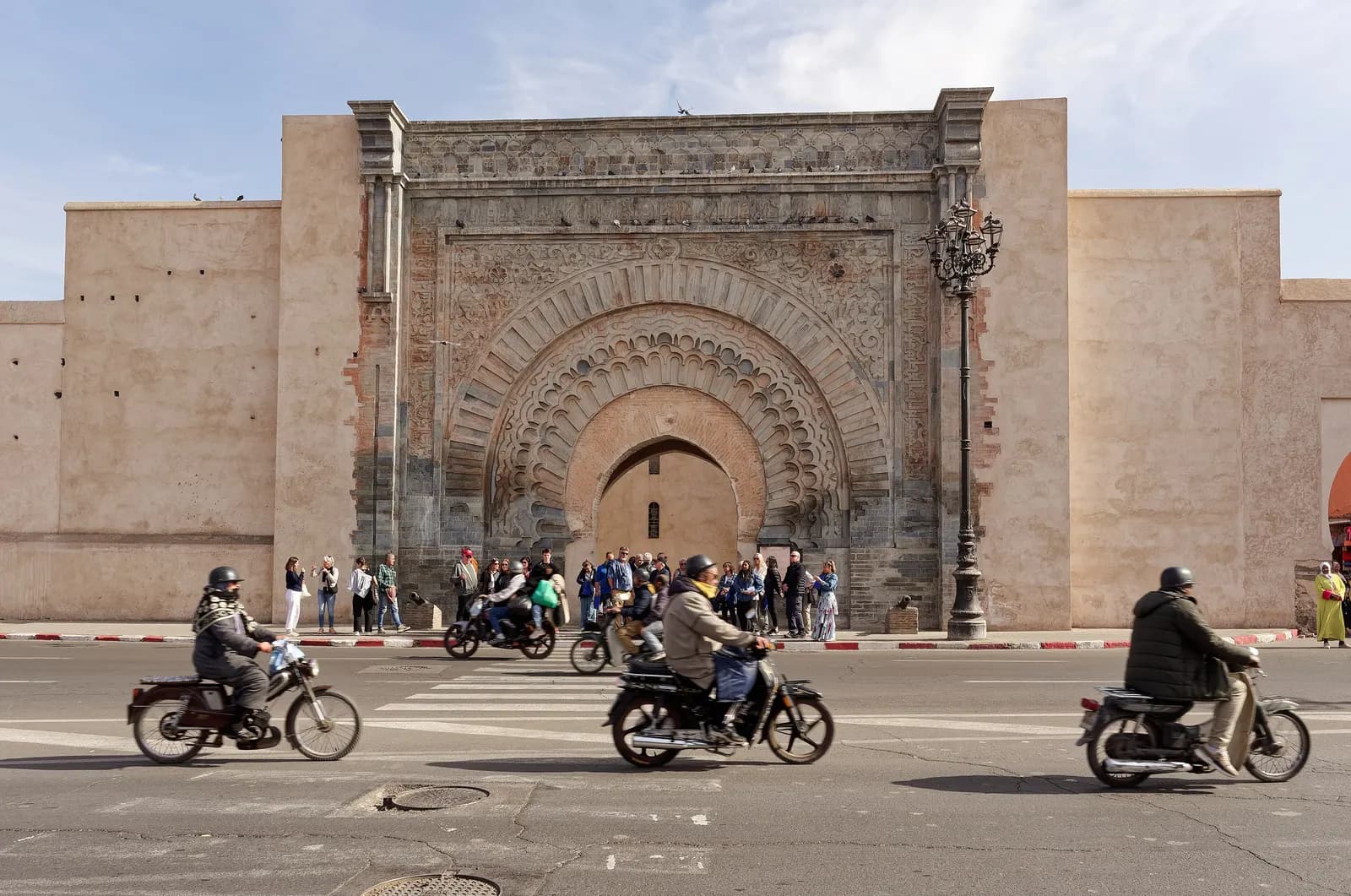 Bab Agnaou, the carved stone gate in the Kasbah wall, with visitors in front of it and mopeds passing on the road