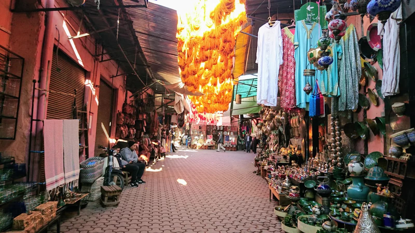 A covered souk lane in the Marrakech medina with pottery, lanterns and robes on display, dyed wool hanging overhead and a moped parked by the wall