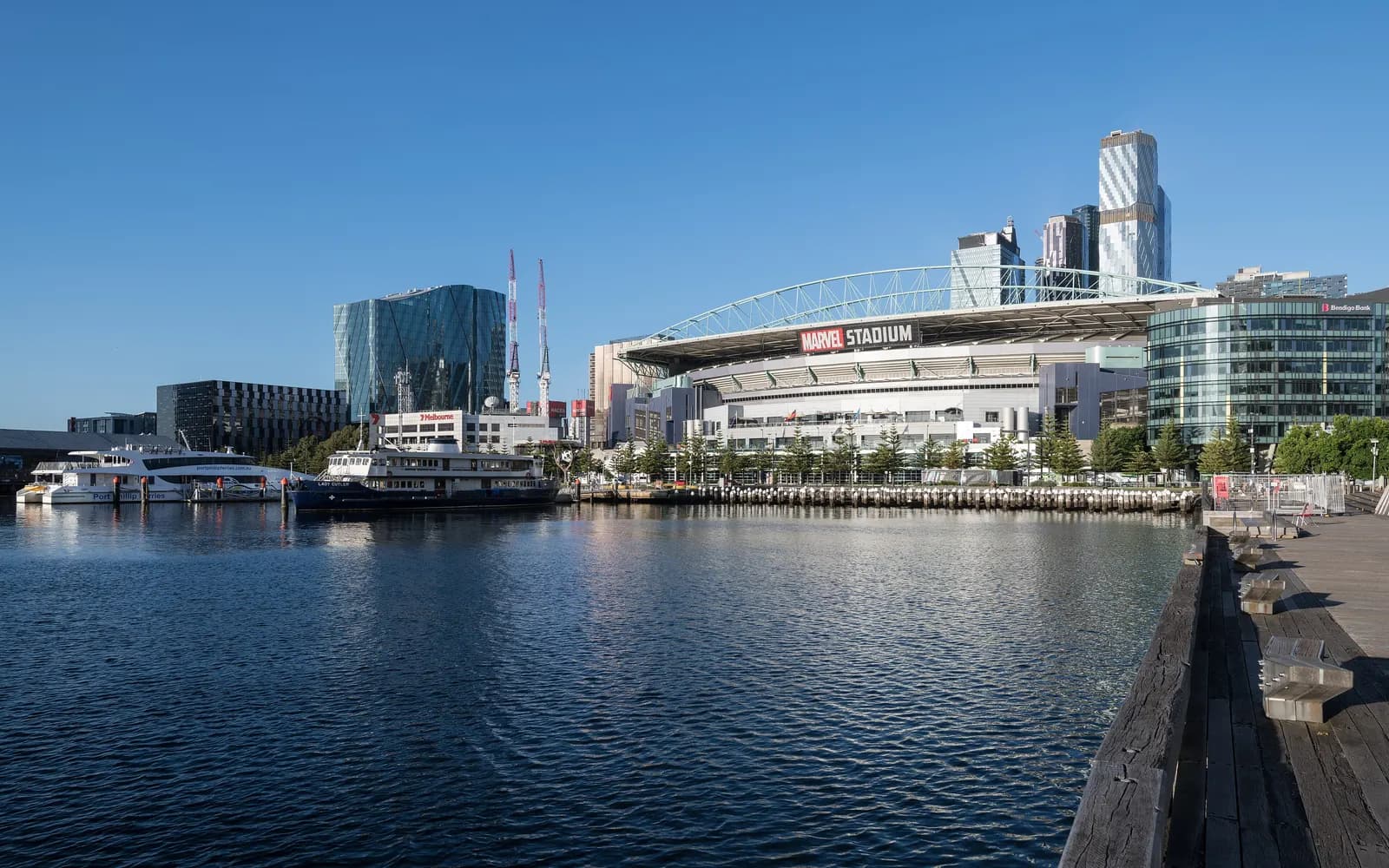 Victoria Harbour in Docklands, Melbourne, with ferries moored at a wharf, the Docklands stadium and glass office towers behind and a timber boardwalk on the right
