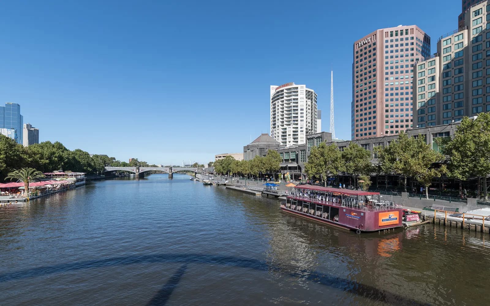 The Yarra River beside Southbank in Melbourne, with the Southgate buildings, riverside trees and a moored restaurant boat on the right bank and a bridge in the distance