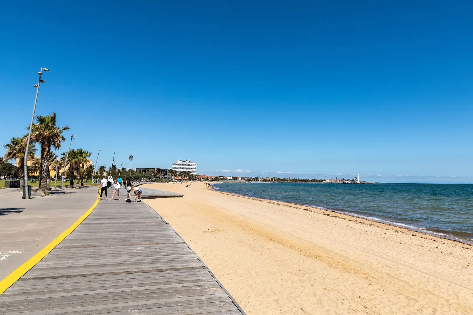 St Kilda Beach in Melbourne, with a timber boardwalk, palm trees and street lights along the foreshore path, the sand and the water of Port Phillip Bay