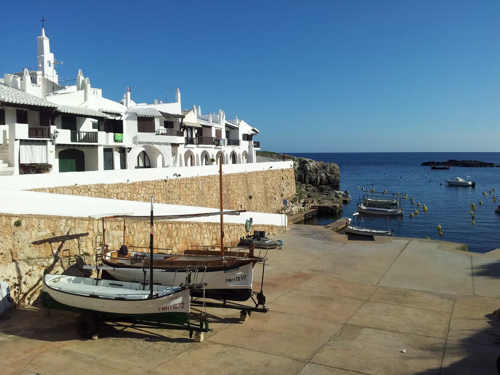White houses with a small belfry above the slipway at Caló d'en Fus, next to Binibèquer Vell, with boats on trailers and the sea beyond.