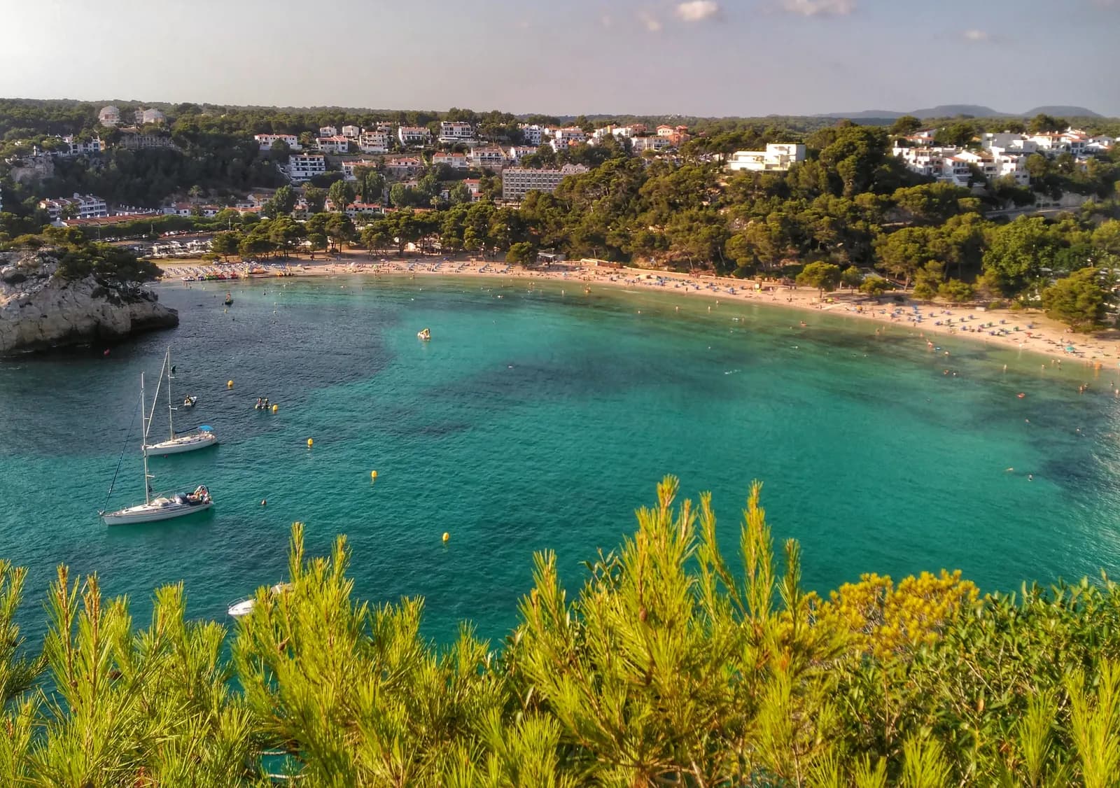 The horseshoe bay of Cala Galdana seen from the cliff, with turquoise water, anchored sailing boats and hotels among the pines.