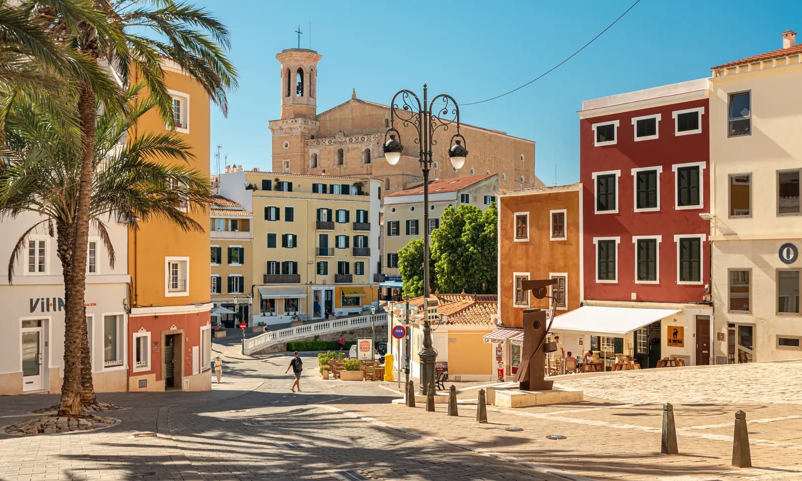 A square in the centre of Maó with coloured town houses, a palm tree and a church bell tower behind.