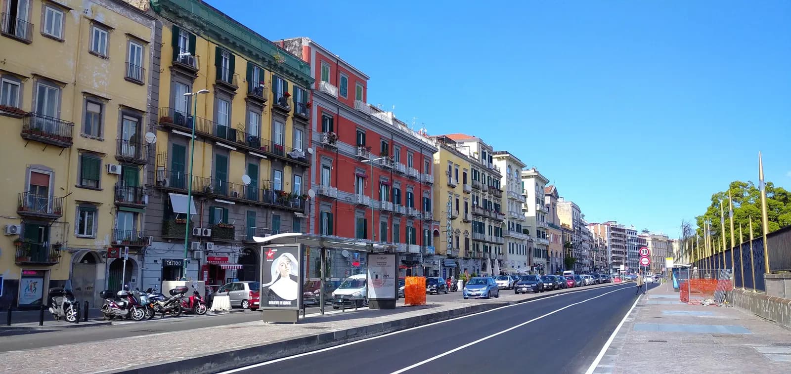 The Riviera di Chiaia with yellow, green and red apartment buildings, a bus shelter and parked scooters under a blue sky.