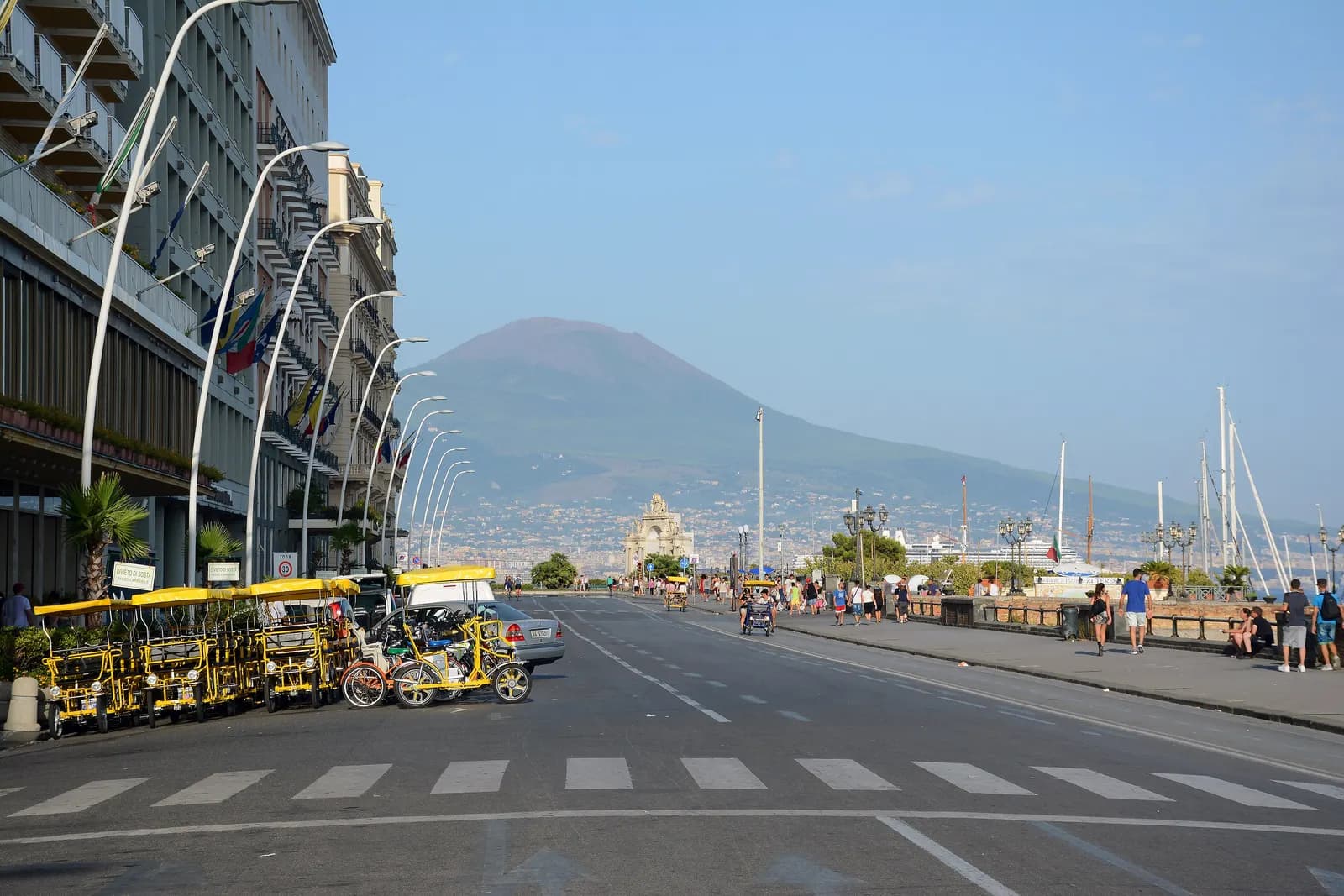 Via Partenope on the Naples seafront, with hotels, a row of pedal cars, walkers and Vesuvius in the haze.