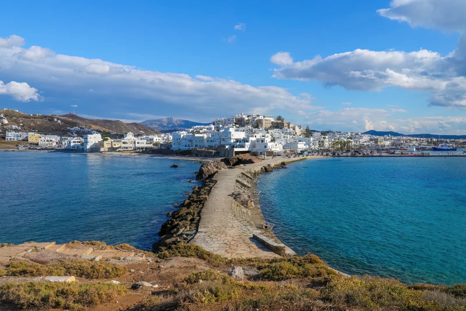 Naxos town seen from the islet of Palatia across the stone causeway, with the white houses of Chora rising to the Kastro and mountains behind
