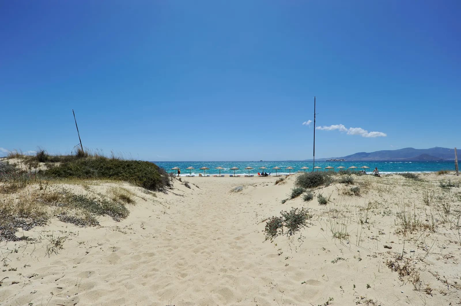 A sandy path through the dunes to Plaka beach, with a row of straw umbrellas at the water's edge