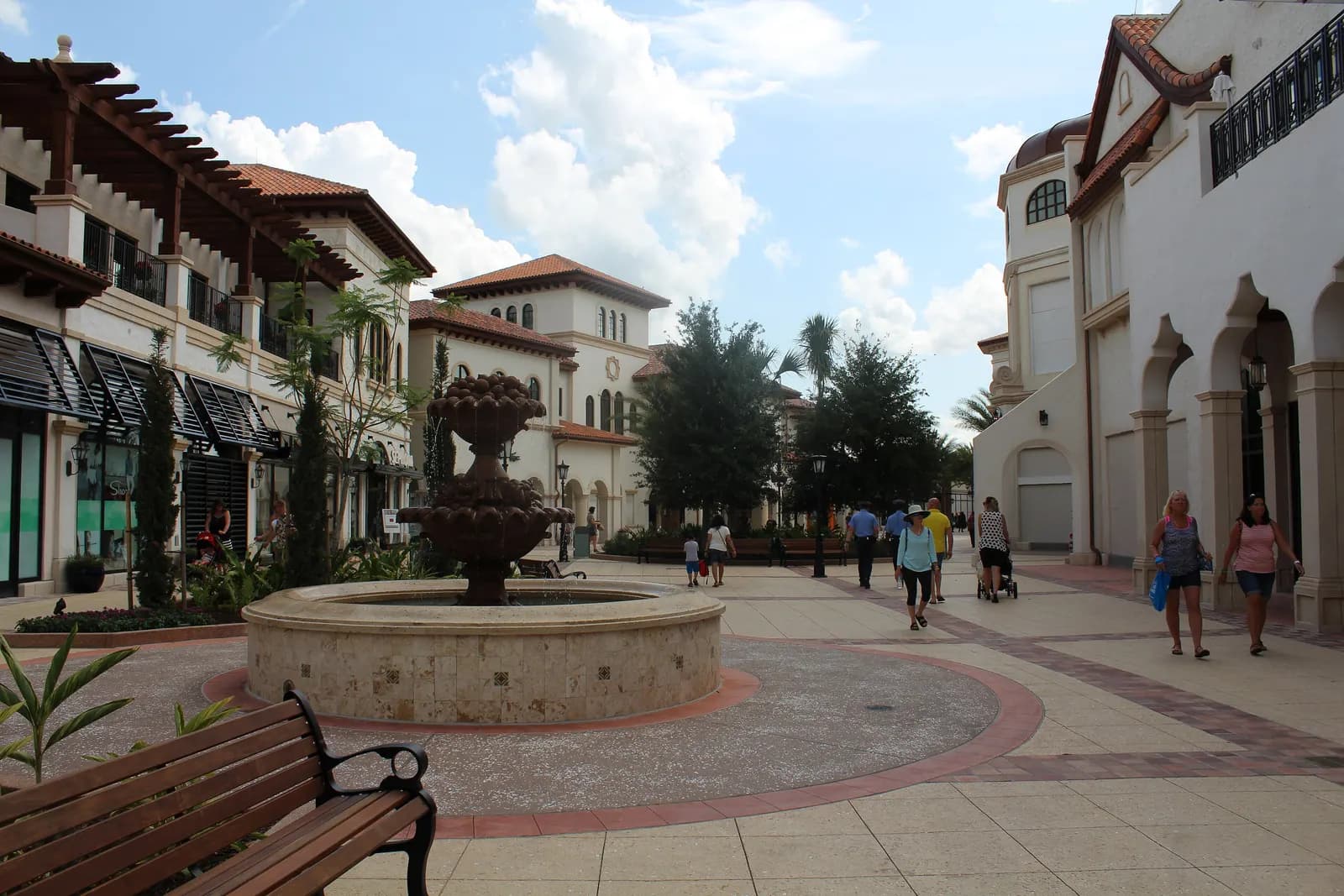 A fountain in a paved plaza between cream Spanish-style shop buildings with tiled roofs at the Town Center of Disney Springs, with shoppers walking under a partly cloudy sky