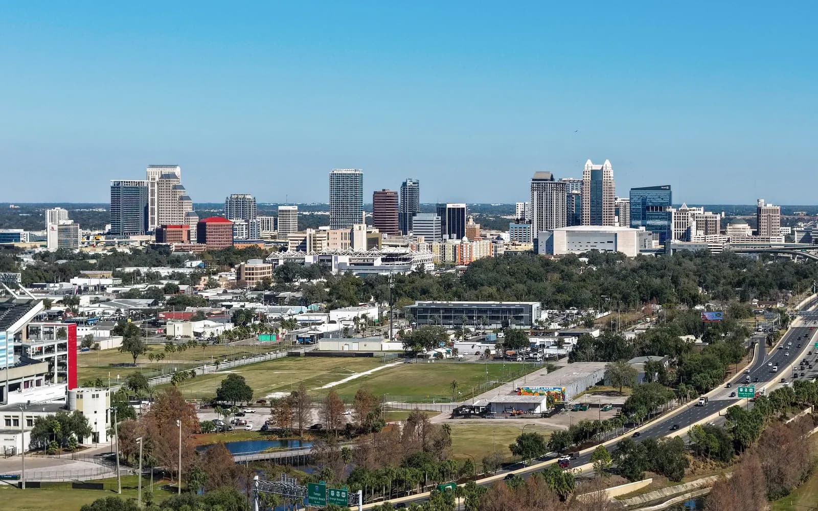 The downtown Orlando skyline seen from the air on a clear day, with an elevated highway on the right and low buildings, trees and open land in front