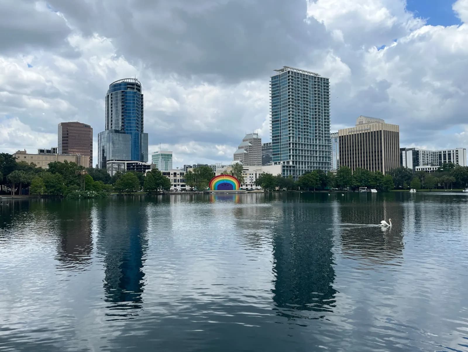 Downtown Orlando towers behind Lake Eola, with a rainbow-coloured bandshell on the far shore, a swan on the water and a cloudy sky reflected in the lake