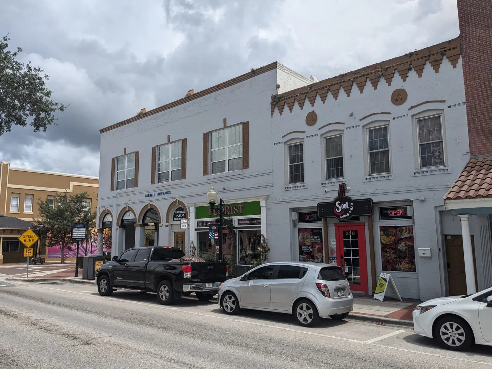 Two-storey white brick commercial buildings with a florist, offices and a restaurant on Broadway in downtown Kissimmee, with cars parked along the street under a cloudy sky