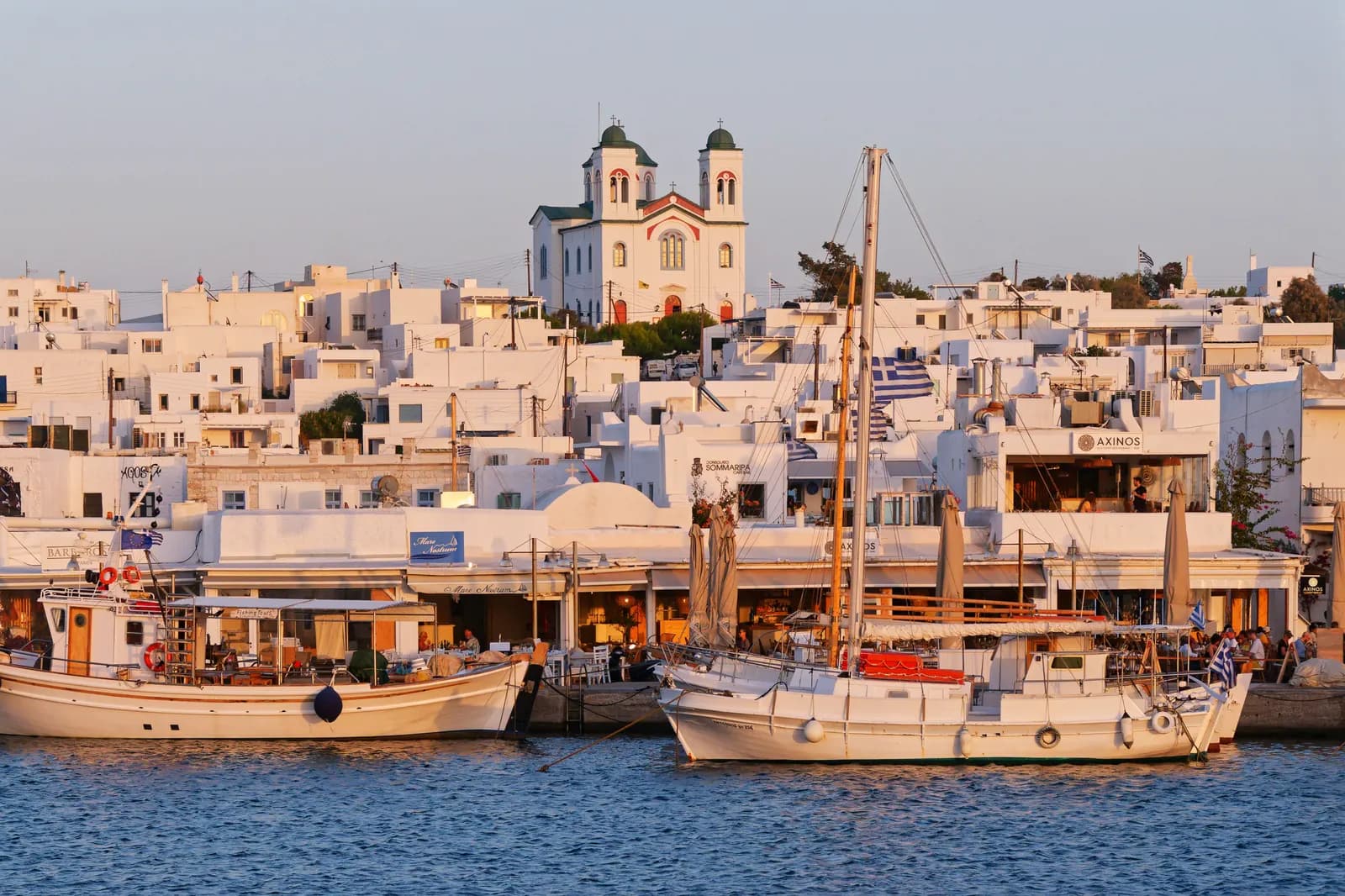 Fishing boats moored along the quay of Naousa harbour in front of waterfront tavernas, with white houses and a twin-towered church above