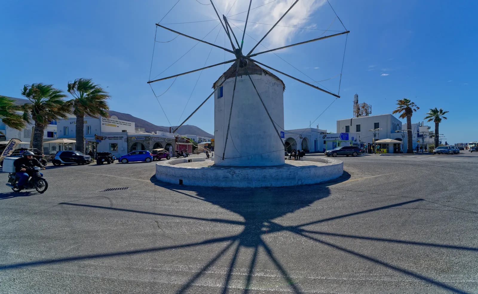 The whitewashed windmill on the roundabout by Parikia port, with palm trees, parked cars and shops around it