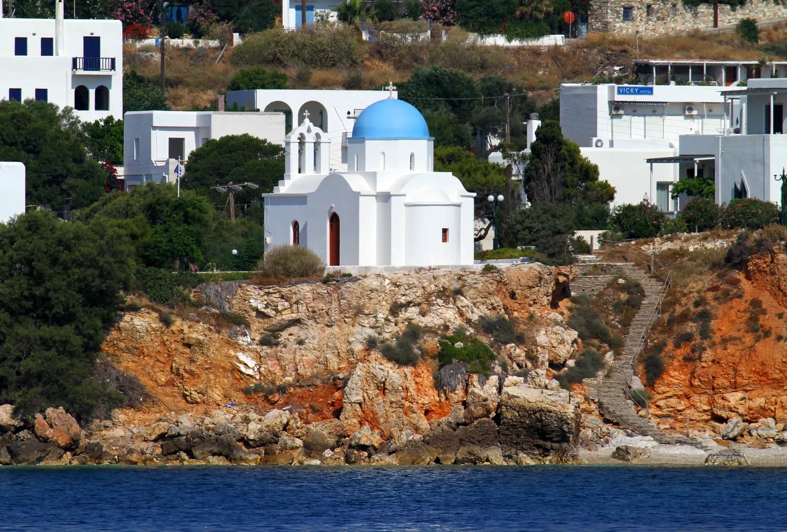 A white chapel with a blue dome on the red rocks above the sea at Piso Livadi, with houses and a hotel behind