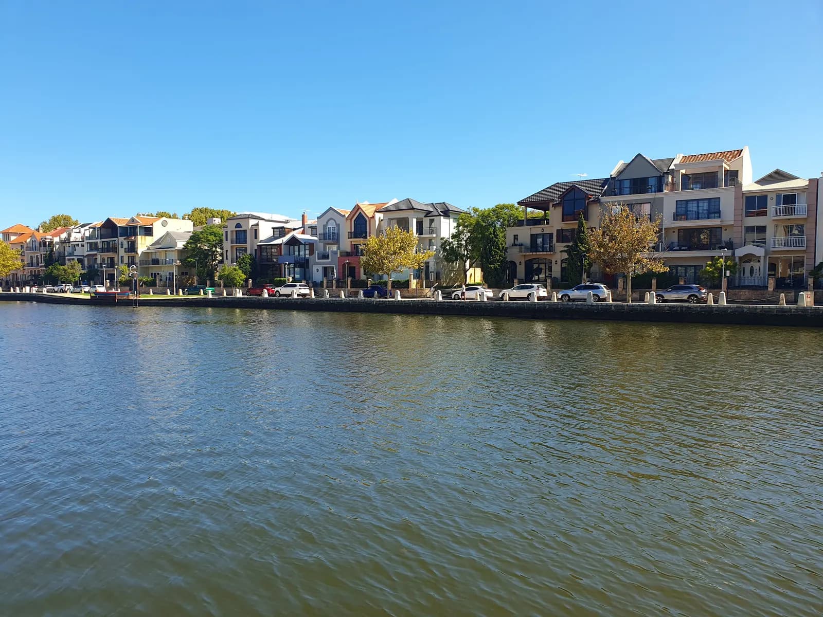 Townhouses and apartment buildings along the water of Claisebrook Cove in East Perth, with cars parked on the waterfront street
