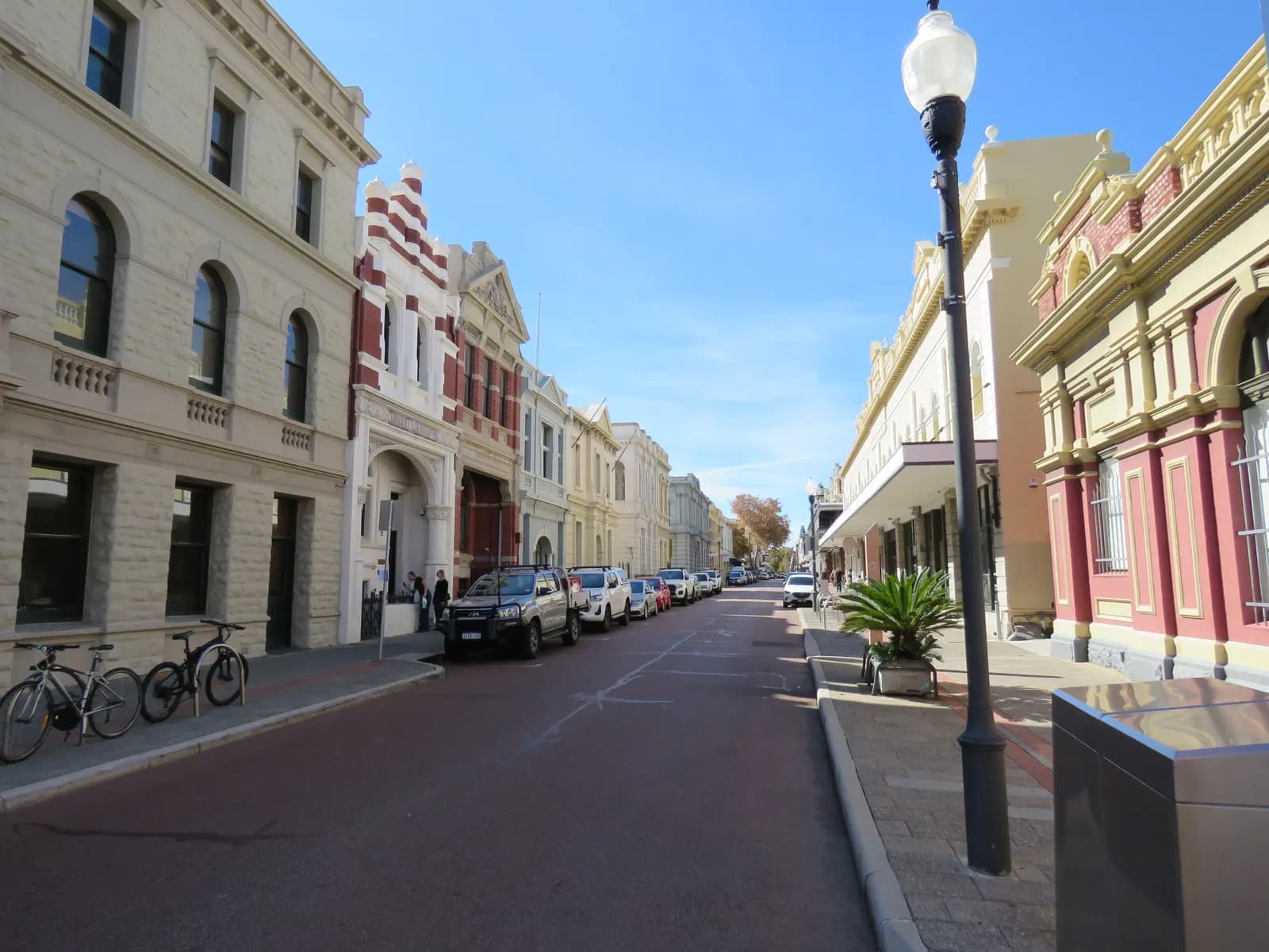 High Street in Fremantle, lined with stone and painted brick buildings with decorated facades, cars parked along one side and bicycles at a rack