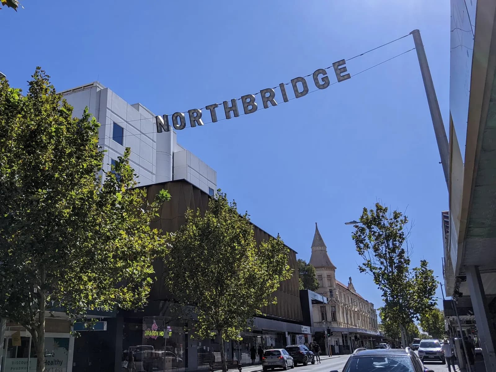 A NORTHBRIDGE sign strung across a street in Northbridge lined with trees and shops, with cars on the road and a building with a spire further along