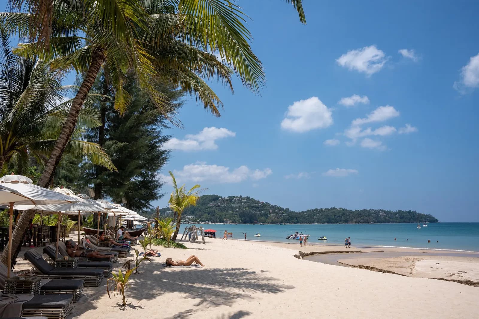 Sun loungers and white umbrellas under palm trees on Bang Tao Beach, with pale sand, jet skis in the bay and a wooded headland under a blue sky