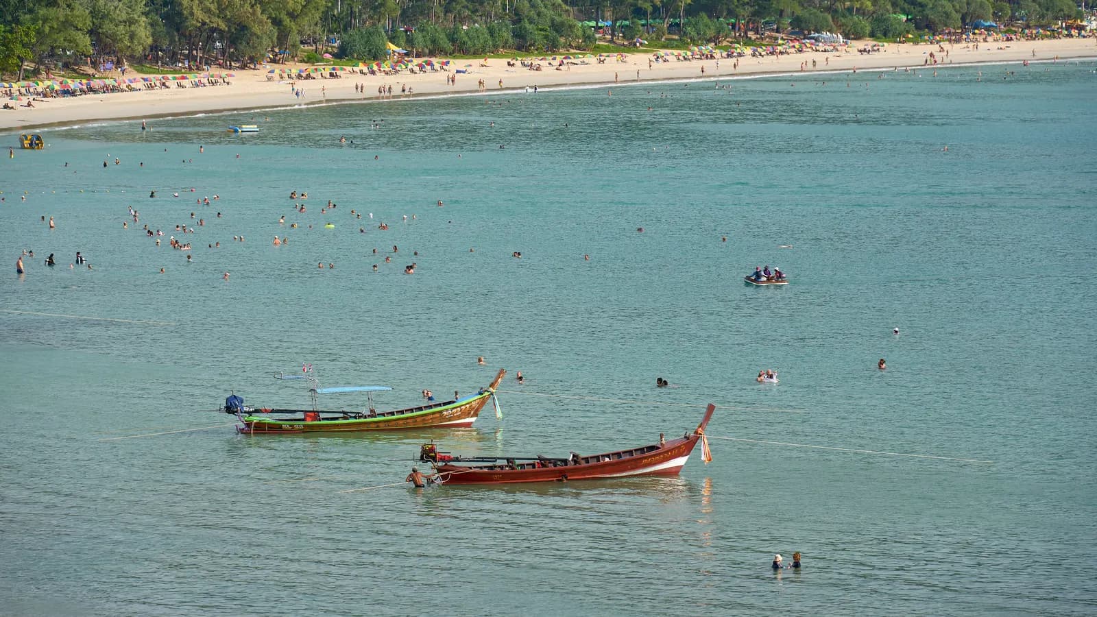 Longtail boats and swimmers in the turquoise water of Kata Beach, with sunshades along the sand and trees behind