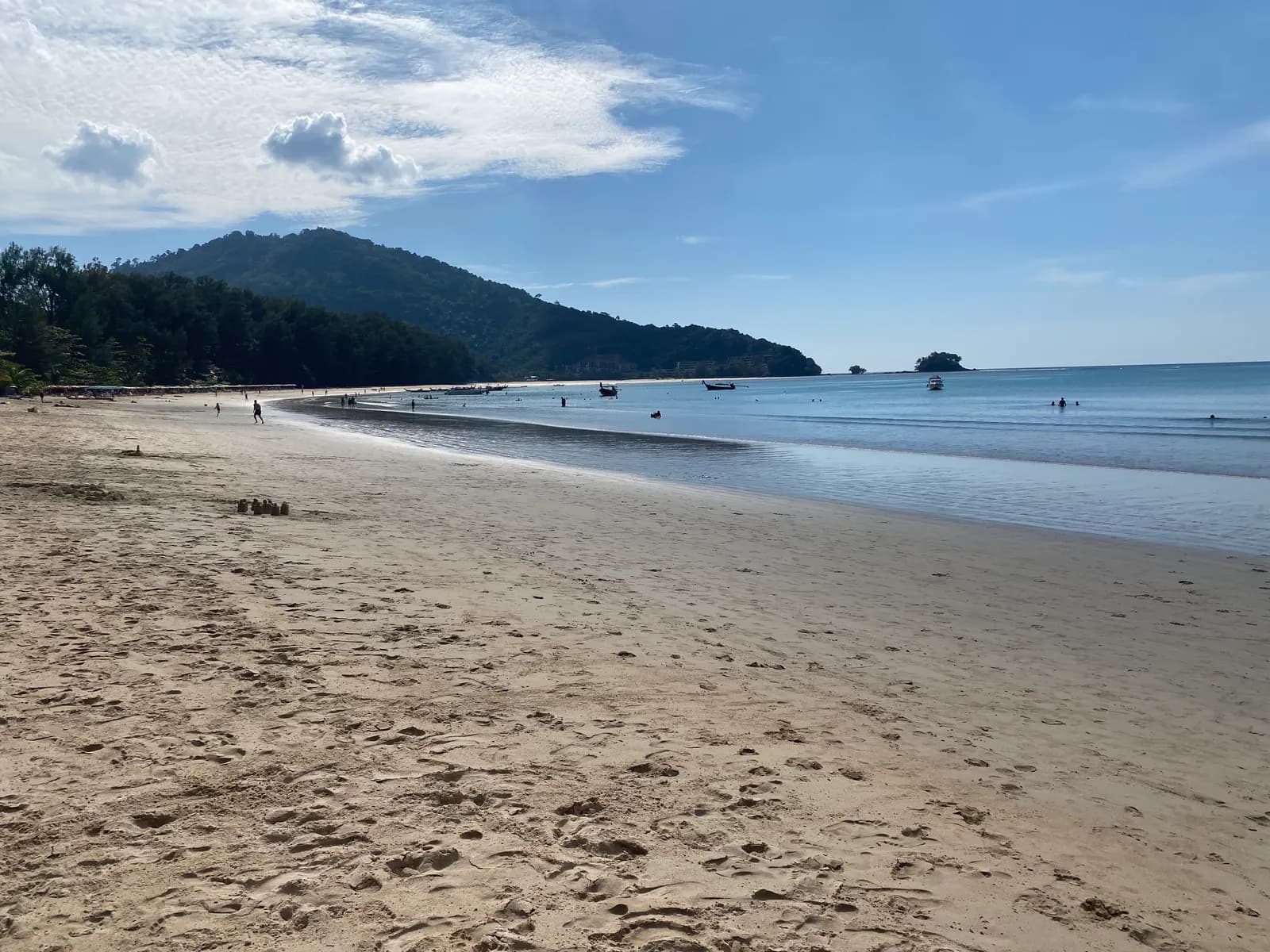 The long sandy curve of Nai Yang Beach with longtail boats in the calm bay, a forested hill behind and a small island offshore
