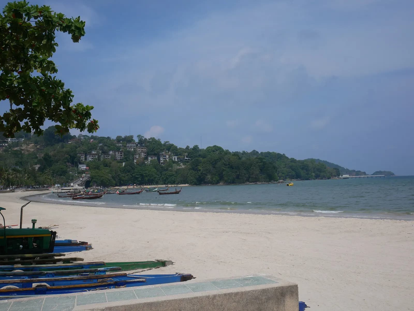 The south end of Patong Beach, with longtail boats pulled up on the white sand and moored in the bay below a wooded headland with hillside buildings