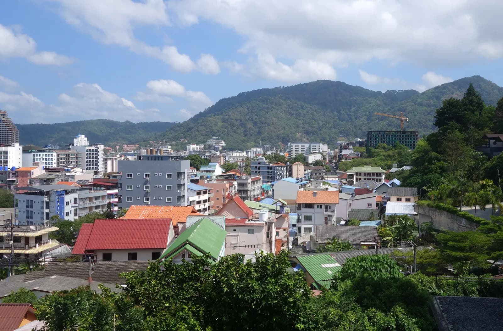 Rooftops and hotel blocks of Patong town filling the valley below green hills, with a crane on a new building