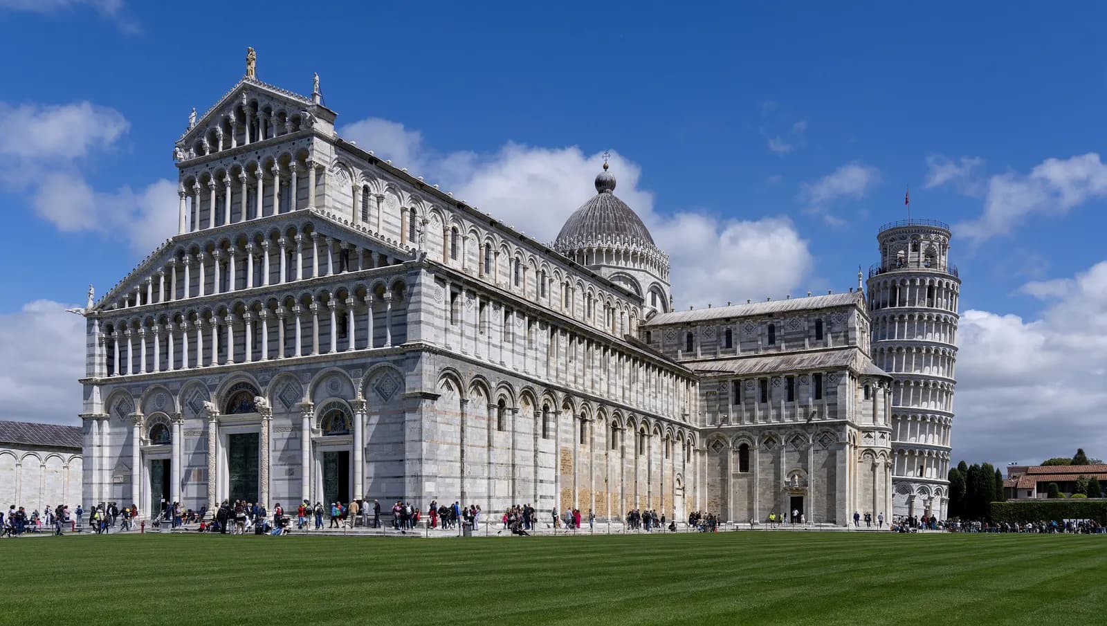 Pisa cathedral and the Leaning Tower on the lawn of the Piazza dei Miracoli, under a blue sky with clouds.