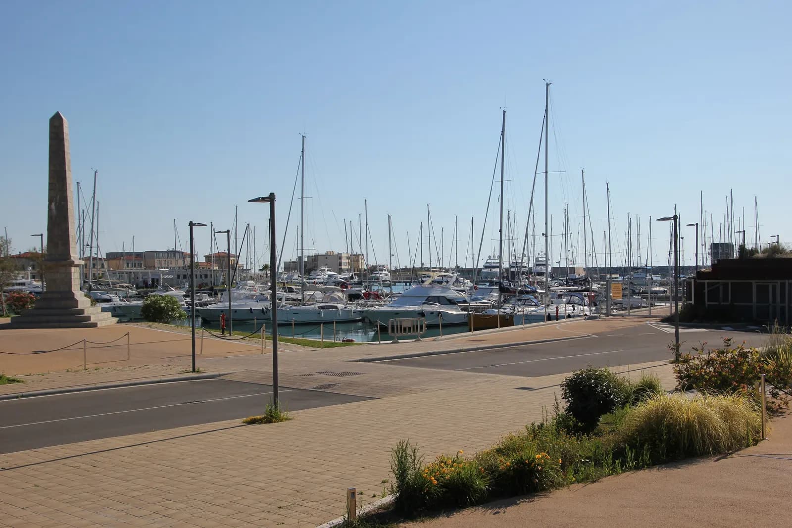 The Marina di Pisa harbour, with sailing boats and motor yachts moored beyond a stone obelisk and a paved promenade.