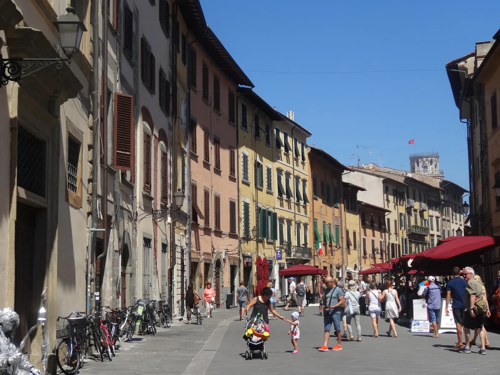 Via Santa Maria in Pisa on a summer day, with shuttered houses, café awnings and the top of the Leaning Tower at the end of the street.
