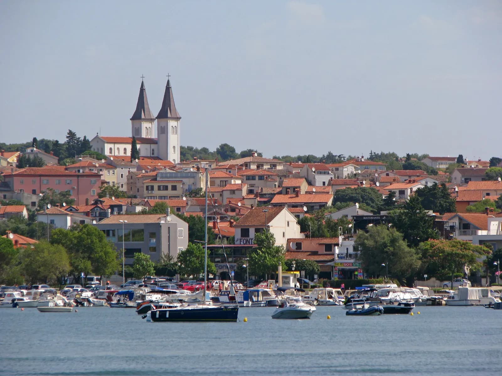 Medulin seen across its harbour, with small boats, a waterfront car park and the twin towers of the parish church above the houses