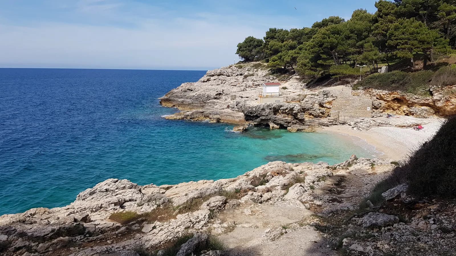 A small pebble cove on the rocky Verudela shore in Pula, with pine trees above the beach and open sea beyond