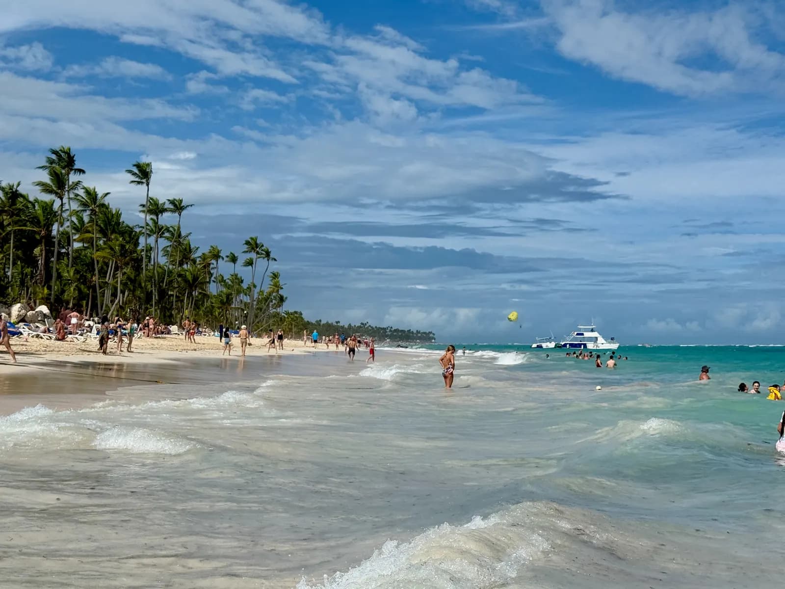 Bávaro beach with swimmers in the turquoise surf, people walking on the sand, a line of palm trees and a boat offshore