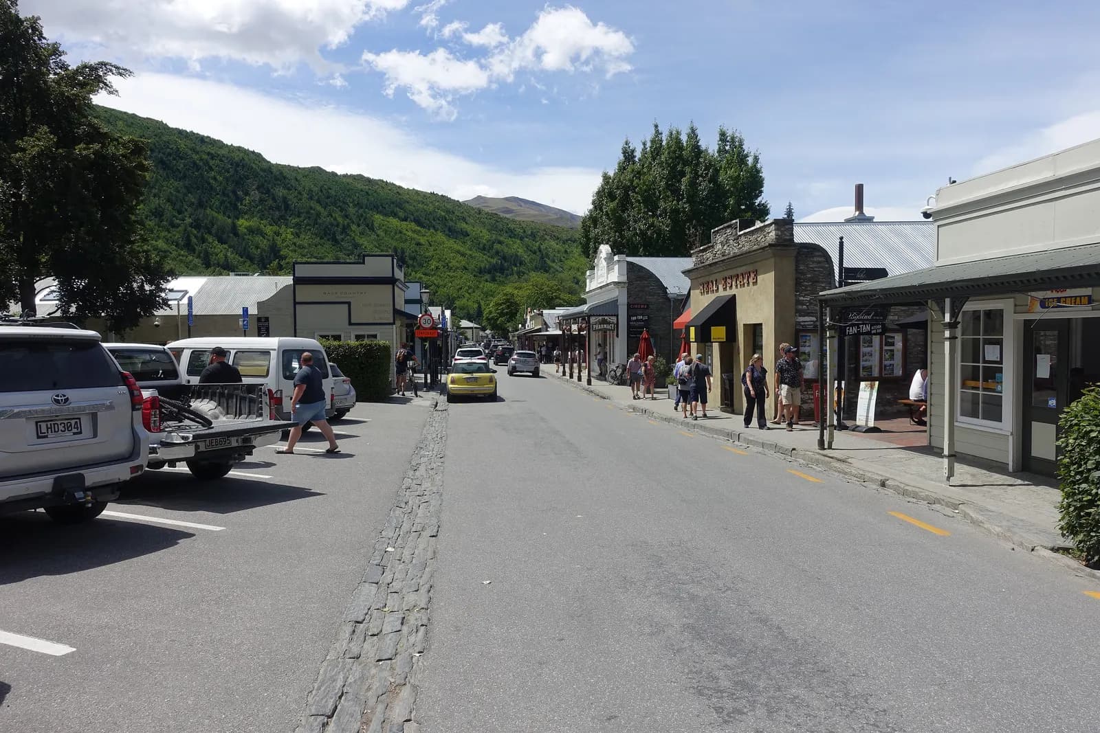 Buckingham Street in Arrowtown with cars parked along the kerb, people on the footpath outside old shopfronts and wooded hills behind