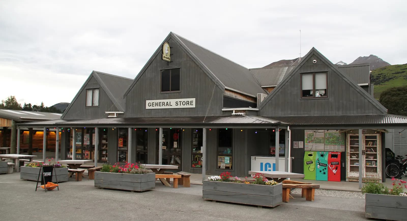 The dark wooden General Store in Glenorchy, with picnic tables and flower planters in front and a hill behind
