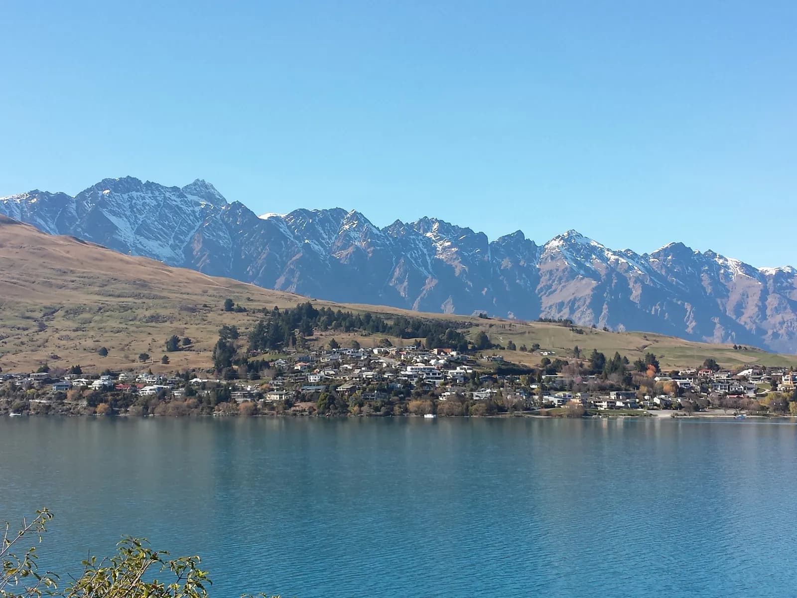 Houses of Kelvin Heights along the shore of Lake Wakatipu, with the snow-streaked Remarkables range behind under a clear sky