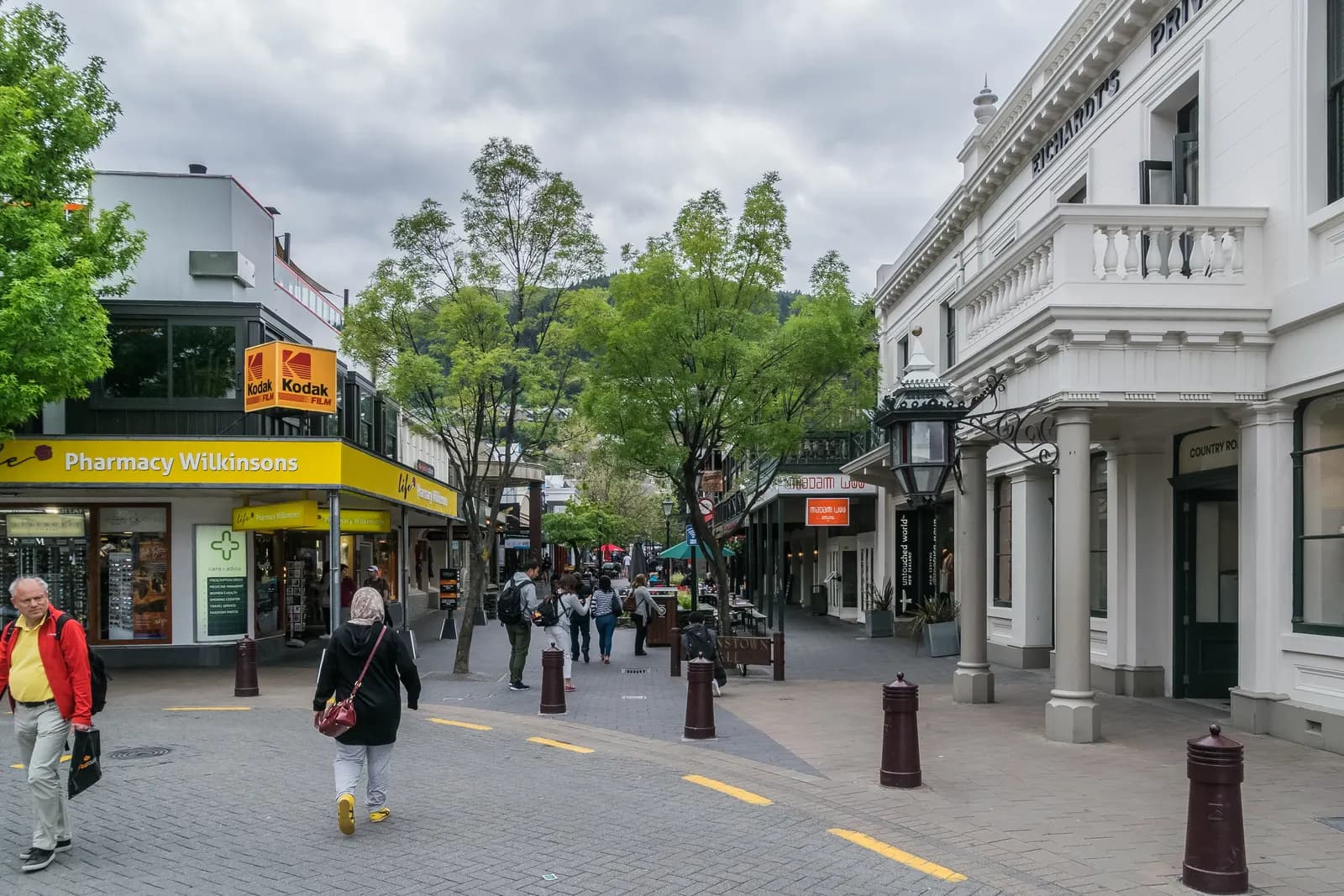 The pedestrian Queenstown Mall with shops, street lamps and trees, and people walking under a cloudy sky