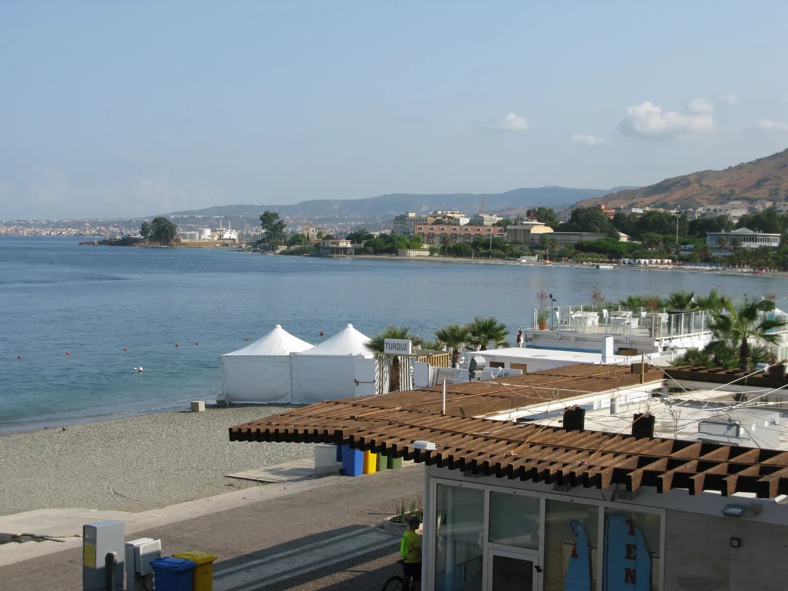 Beach clubs with white marquees on the shore below Reggio Calabria's Lungomare Falcomata, with the bay curving north towards low hills.